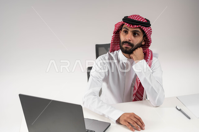 تعابير التفكير في استراتيجيات وأهداف العمل، بورتريه مقرب لموظف عربي سعودي خليجي يرتدي الشماغ والثوب التقليدي يقوم بالتخطيط والتأمل داخل مكتبه, إيماءات التركيز والتمعن, إدارة شؤون الأعمال وتنظيمها, النظر إلى شيء ما في الأعلى, خلفية بيضاء .