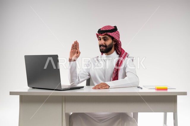مكالمات فيديو وبث مباشر, التلويح باليد والسلام عن بعد، بورتريه لرجل عربي سعودي خليجي مبتسم يرتدي الشماغ والثوب التقليدي يقوم باستخدام جهاز الحاسوب المحمول, التواصل مع الأهل والأصدقاء, اجتماعات عمل أونلاين, خلفية بيضاء .