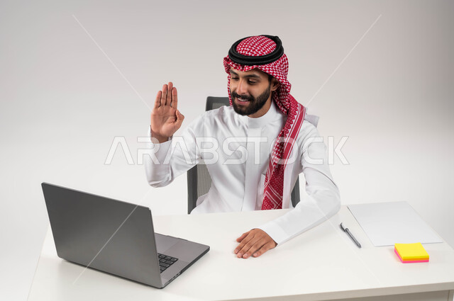 Waving a hand and saying hello from a distance, a close-up portrait of a smiling Saudi Gulf Arab man wearing a shemagh and traditional dress, using a laptop, video calls and live broadcasts, communicating with family and friends, online business meetings, white background