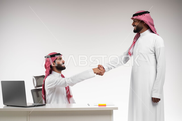 الاتفاق لتوقيع صفقات شراكة جديدة، مفهوم الإدارة المتعاونة، بورتريه لرجلين عربيين خليجيين سعوديين مبتسمين يرتديان الشماغ والثوب التقليدي يصافحان بعضهما بإيماءات السرور، الوحدة والتفاعل الجماعي لتحقيق أهداف الشركة وحل المشكلات، خلفية بيضاء.