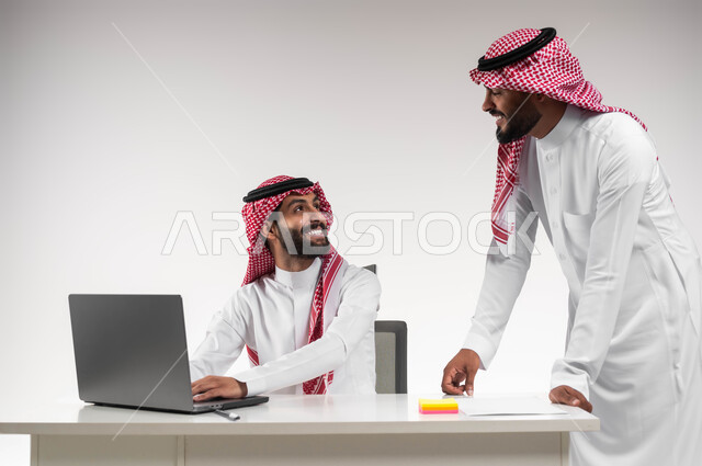 الوحدة والتفاعل الجماعي لتحقيق أهداف الشركة وحل المشكلات، مفهوم الإدارة المتعاونة، بورتريه لرجلان عربيان خليجيان سعوديان مبتسمان يرتديان الشماغ والثوب التقليدي يناقشان في أمور العمل باستخدام الحاسوب المحمول، إنجاز المهام المكتبية اليومية، خلفية بيضاء.