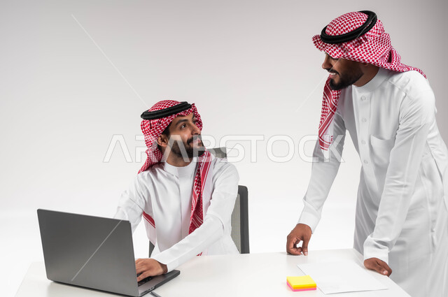 مفهوم الإدارة المتعاونة، بورتريه لموظفان عربيان خليجيان سعوديان مبتسمان يرتديان الشماغ والثوب التقليدي يناقشان في أمور العمل باستخدام الحاسوب المحمول، إنجاز المهام المكتبية اليومية، الوحدة والتفاعل الجماعي لتحقيق أهداف الشركة وحل المشكلات، خلفية بيضاء.