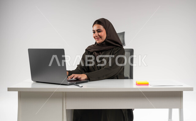 Communicating with customers through modern technical devices, completing office tasks with love and passion, portrait of a Saudi Gulf Arab businesswoman wearing an abaya, sitting behind the table and working on a laptop, achieving the company’s goals, a successful and comfortable work environment, white background