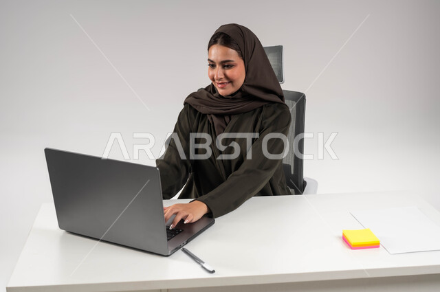 انجاز المهام المكتبيه بحب وشغف، بورتريه لسيدة أعمال عربية خليجية سعودية ترتدي العباءة والحجاب تقوم بالعمل على جهاز الحاسوب المحمول، تحقيق أهداف الشركة، بيئة عمل ناجحة ومريحة، التواصل مع العملاء من خلال الاجهزة التقنية الحديثة، خلفية بيضاء