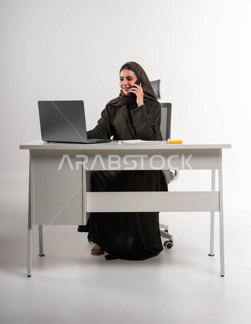 التواصل مع الموظفين عبر الجوال، دمج التقنية والتكنولوجيا في الحياة العملية، إجراء مكالمة عمل في مقر الشركة، بورتريه لسيدة أعمال عربية خليجية سعودية مبتسمة ترتدي العباءة والحجاب تجلس وراء الطاولة وتعمل على الحاسوب المحمول، إنجاز المهام المكتبية بحب وشغف، خلفية بيضاء