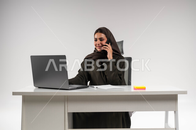 دمج التقنية والتكنولوجيا في الحياة العملية، إجراء مكالمة عمل في مقر الشركة، بورتريه لسيدة أعمال عربية خليجية سعودية مبتسمة محجبة ترتدي العباءة السوداء تعمل على الحاسوب المحمول ،إنجاز المهام المكتبية بحب وشغف، التواصل مع الموظفين عبر الجوال، خلفية بيضاء