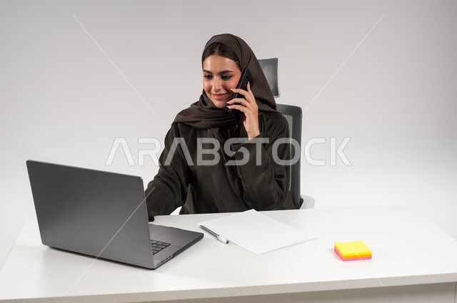 إجراء مكالمة عمل في مقر الشركة، بورتريه مقرب لسيدة أعمال عربية خليجية سعودية مبتسمة محجبة ترتدي العباءة السوداء تعمل على الحاسوب المحمول ،دمج التقنية والتكنولوجيا في الحياة العملية، إنجاز المهام المكتبية بحب وشغف، التواصل مع الموظفين عبر الجوال، خلفية بيضاء