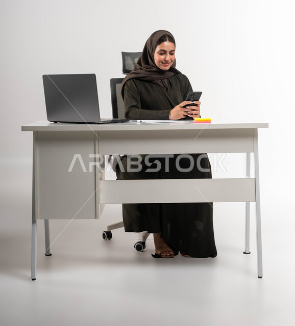 الدردشة مع الأهل والأصدقاء، دمج التقنية و التكنولوجية في الحياة العملية، التسوق الالكتروني عبر برامج الهاتف المحمول، بورتريه لامرأة عربية خليجية سعودية محجبة ترتدي العباءة السوداء و تحمل بيدها الجوال بإيماءات السرور، خلفية بيضاء