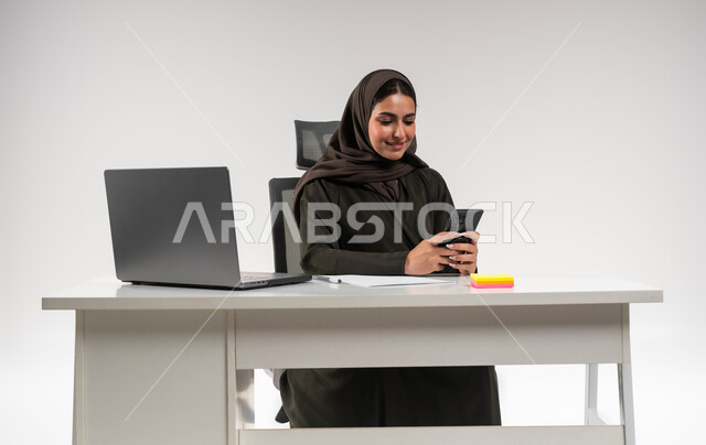 دمج التقنية و التكنولوجية في الحياة العملية، التسوق الالكتروني عبر برامج الهاتف المحمول، الدردشة مع الأهل والأصدقاء، بورتريه لامرأة عربية خليجية سعودية محجبة ترتدي العباءة السوداء و تحمل بيدها الجوال بإيماءات السرور، خلفية بيضاء