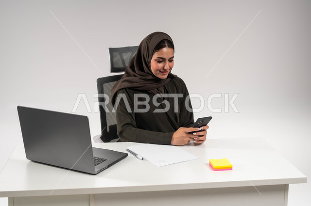 التسوق الالكتروني عبر برامج الهاتف المحمول، الدردشة مع الأهل والأصدقاء، بورتريه مقرب لامرأة عربية خليجية سعودية محجبة ترتدي العباءة تجلس وراء الطاولة و تحمل بيدها الجوال بإيماءات السرور، دمج التقنية و التكنولوجية في الحياة العملية ،خلفية بيضاء