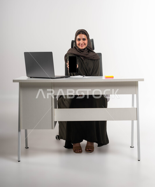 شركات ومكاتب سعودية، عرض التطبيقات والبرامج الخاصة بالاعمال التجارية عبر الهاتف المحمول، بورتريه لامرأة عربية خليجية سعودية محجبة ترتدي العباءة السوداء تحمل بيدها الجوال بشاشة سوداء فارغه  بإيماءات السرور، دمج التقنية و التكنولوجية في الحياة العملية ،خلفية بيضاء