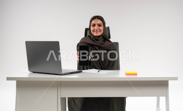 عرض التطبيقات والبرامج الخاصة بالاعمال التجارية عبر الهاتف المحمول، بورتريه لامرأة عربية خليجية سعودية محجبة ترتدي العباءة تجلس وراء الطاولة و تحمل بيدها الجوال بشاشة سوداء فارغة، دمج التقنية و التكنولوجية في الحياة العملية، شركات ومكاتب سعودية، خلفية بيضاء