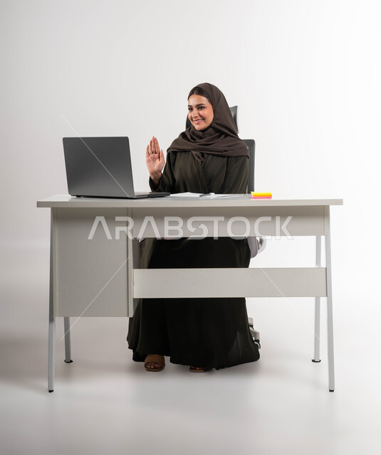 اجتماعات عمل أونلاين, القاء التحية والسلام عن بعد، مكالمات فيديو وبث مباشر, بورتريه لامرأة عربية خليجية سعودية مبتسمة محجبة ترتدي العباءة تجلس وراء الطاولة ترفع يدها للتلويح وتقوم باستخدام جهاز الحاسوب, التواصل مع الأهل والأصدقاء, خلفية بيضاء