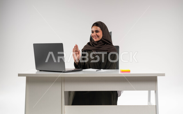 القاء التحية والسلام عن بعد، مكالمات فيديو وبث مباشر, بورتريه لسيدة أعمال عربية خليجية سعودية مبتسمة محجبة ترتدي العباءة تجلس وراء الطاولة ترفع يدها للتلويح و تقوم باستخدام جهاز الحاسوب المحمول, التواصل مع الأهل والأصدقاء, اجتماعات عمل أونلاين, خلفية بيضاء