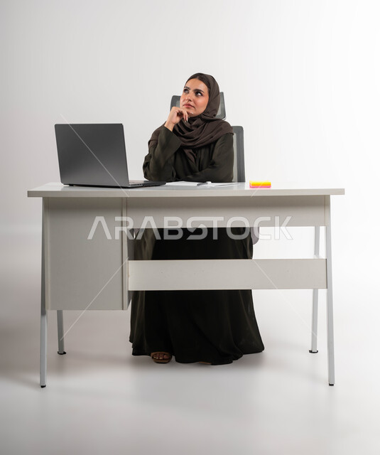 Thinking about developing new plans for business projects, portrait of a veiled Saudi Arabian Gulf woman wearing an abaya, sitting behind the table and placing her hand on her face with gestures, finding a solution to a problem, using modern technologies, deducing ideas to achieve success and the company’s goals, white background.