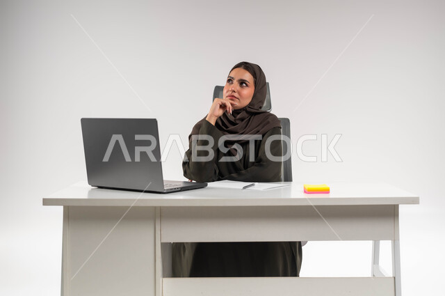 Looking up and thinking about developing new plans for business projects, deducing ideas to achieve success and the company’s goals, portrait of a veiled Saudi Arabian Gulf businesswoman wearing an abaya sitting behind the table and placing her hand on her face with gestures of finding a solution to a problem, white background