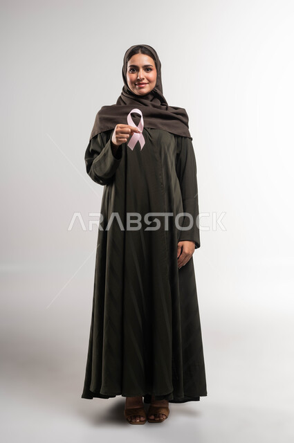 الكشف المبكر عن المرض والاهتمام بعلاجه ،رمز الحملة العالمية، بورتريه لامرأة عربية خليجية سعودية محجبة ترتدي العباءة وتحمل على كفيها شريط زهري اللون، اليوم العالمي للتوعية بسرطان الثدي، النظر إلى الكاميرا بإيماءات السرور، صورة الجسم بالطول الكامل، خلفية بيضاء
