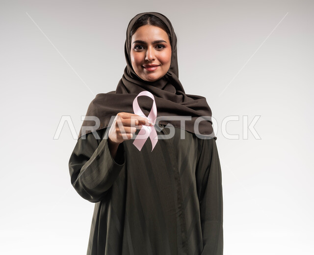 النظر إلى الكاميرا بإيماءات السرور، اليوم العالمي للتوعية بسرطان الثدي، رمز الحملة العالمية، بورتريه مقرب لامرأة عربية خليجية سعودية محجبة مبتسمة ترتدي العباءة وتحمل بيدها شريط زهري اللون، الاهتمام بالمرض و بالكشف المبكر ،خلفية بيضاء