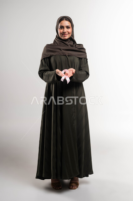 رمز الحملة العالمية، بورتريه لامرأة عربية خليجية سعودية محجبة ترتدي العباءة وتحمل على كفيها شريط زهري اللون،  اليوم العالمي للتوعية بسرطان الثدي، الاهتمام بالمرض و بالكشف المبكر ،النظر إلى الكاميرا بإيماءات السرور، صورة الجسم بالطول الكامل، خلفية بيضاء