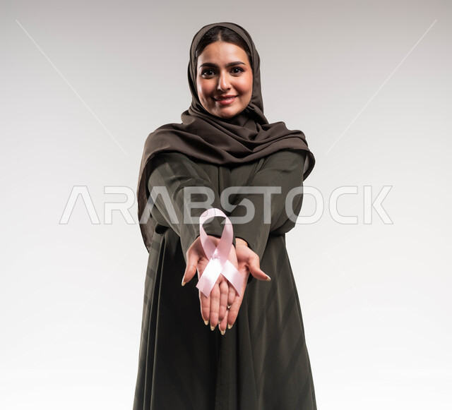رمز الحملة العالمية، حملة توعوية للكشف المبكر عن سرطان الثدي، بورتريه مقرب لإمرأة عربية خليجي سعودية ترتدي الحجاب والعباءة السوداء تحمل بيديها الشريط الوردي تنظر الى الكاميرا بنظرات القوة، تحدي الأمراض الخبيثة، اليوم العالمي للتوعية والكشف المبكر، خلفية بيضاء