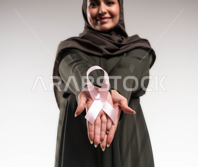 رمز الحملة العالمية، تحدي الأمراض الخبيثة، بورتريه مقرب لإمرأة عربية خليجي سعودية ترتدي الحجاب والعباءة السوداء تحمل بيديها الشريط الوردي تنظر الى الكاميرا بنظرات القوة، حملة توعوية للكشف المبكر عن سرطان الثدي، اليوم العالمي للتوعية والكشف المبكر، خلفية بيضاء