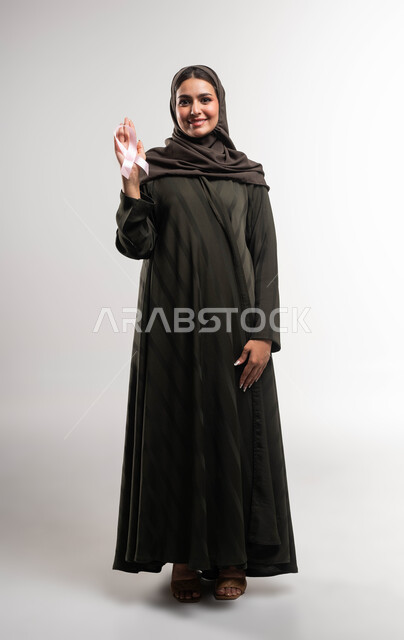 الوقوف بوجه الأمراض الخبيثة، نشر الوعي حول ضرورة الكشف المبكر، حملة نسائية توعوية عن سرطان الثدي، بورتريه لإمرأة عربية خليجي سعودية ترتدي الحجاب والعباءة تضع على يدها الشريط الوردي تنظر الى الكاميرا بقوة، رمز الحملة العالمية، صورة الجسم بالكامل، خلفية بيضاء
