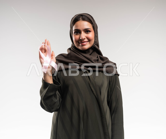 نشر الوعي حول ضرورة الكشف المبكر، حملة نسائية توعوية عن سرطان الثدي، بورتريه مقرب لإمرأة عربية خليجي سعودية مبتسمة ترتدي الحجاب والعباءة السوداء تضع على يدها الشريط الوردي تنظر الى الكاميرا بقوة، تحدي الأمراض والأورام الخبيثة، رمز الحملة العالمية، خلفية بيضاء