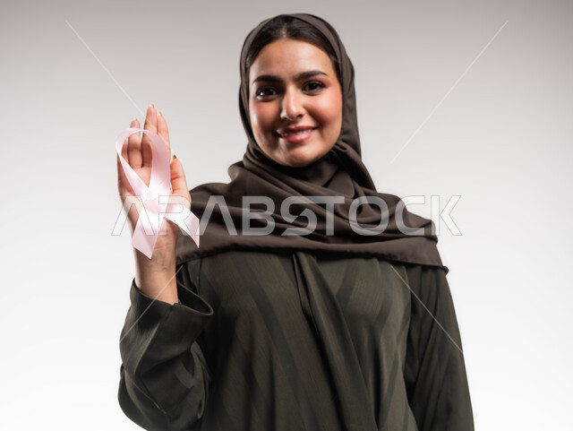 تحدي الأمراض والأورام الخبيثة، نشر الوعي حول ضرورة الكشف المبكر، حملة نسائية توعوية عن سرطان الثدي، بورتريه مقرب لإمرأة عربية خليجي سعودية مبتسمة ترتدي الحجاب والعباءة السوداء تضع على يدها الشريط الوردي تنظر الى الكاميرا بقوة، رمز الحملة العالمية، خلفية بيضاء