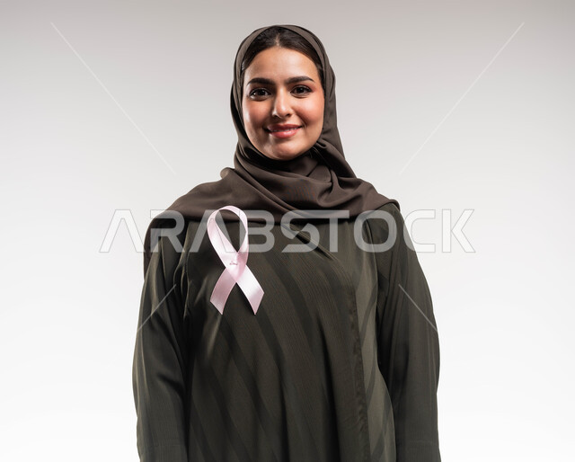 نشر الوعي حول ضرورة الكشف المبكر، حملة نسائية توعوية عن سرطان الثدي، بورتريه مقرب لإمرأة عربية خليجي سعودية مبتسمة ترتدي الحجاب والعباءة السوداء تضع الشريط الوردي تنظر الى الكاميرا بقوة، تحدي الأمراض والأورام الخبيثة، رمز الحملة العالمية، خلفية بيضاء