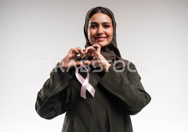 The importance of early detection of breast cancer, a women’s awareness campaign, a close-up portrait of a smiling Saudi Gulf Arab woman wearing the hijab and abaya, wearing a pink ribbon, forming her hands into the shape of a heart, expressions of love and sympathy, the impact of positive support on mental and physical health, white background.