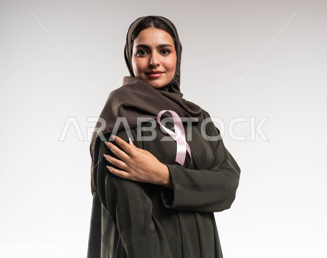 Challenge of malignant diseases, a close-up portrait of a Saudi Arabian Gulf woman wearing the hijab and black abaya, wearing a pink ribbon, looking at the camera with a look of strength, an awareness campaign for early detection of breast cancer, a symbol of the global campaign, World Awareness and Early Detection Day, white background.