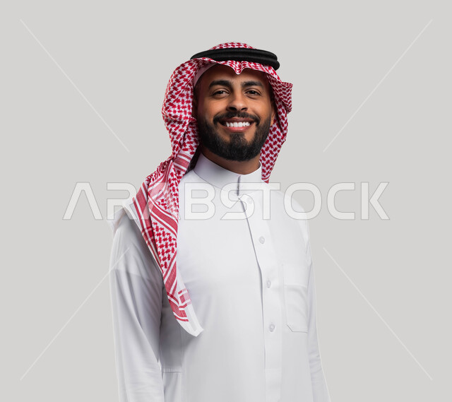 الوقوف بإستقامة وثقة بالنفس، صورة شخصية للوثائق الرسمية، بورتريه مقرب لشاب عربي سعودي خليجي مبتسم يرتدي الشماغ والثوب السعودي التقليدي ينظر إلى الكاميرا بإيماءات السرور والسعادة، خلفية بيضاء