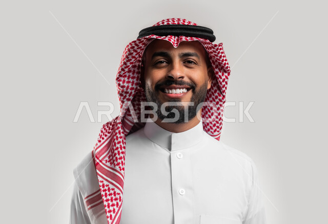 الوقوف بإستقامة وثقة بالنفس, صورة شخصية للوثائق الرسمية، بورتريه مقرب لشاب عربي سعودي خليجي مبتسم يرتدي الشماغ والثوب السعودي التقليدي ينظر إلى الكاميرا بإيماءات السرور والسعادة، خلفية بيضاء