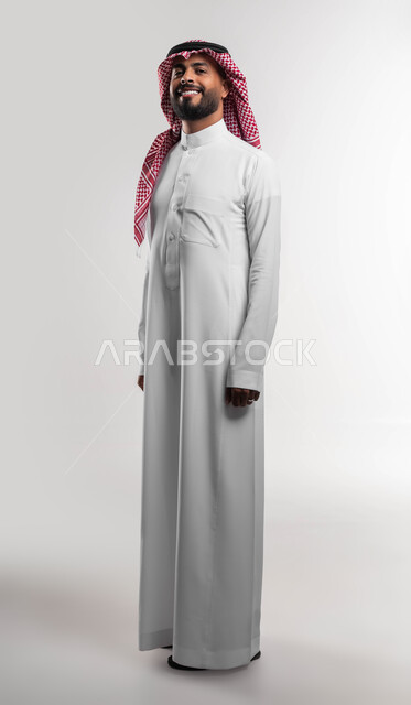 ايماءات الثقة والاعتزاز بالنفس، الاهتمام بالمظهر الخارجي، بورتريه لرجل عربي خليجي سعودي مبتسم يرتدي الشماغ والثوب السعودي التقليدي ينظر إلى الكاميرا بتعابير السرور، مفهوم الاناقه والرجولة، الوقوف باستقامة وانضباط، صورة لكامل الجسم، خلفية بيضاء