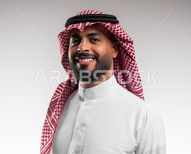 مفهوم الاناقه والرجولة، بورتريه مقرب لرجل عربي خليجي سعودي مبتسم يرتدي الشماغ والثوب السعودي التقليدي ينظر إلى الكاميرا بتعابير السرور، الاهتمام بالمظهر الخارجي، ايماءات الثقة والاعتزاز بالنفس، الوقوف باستقامة وانضباط، خلفية بيضاء