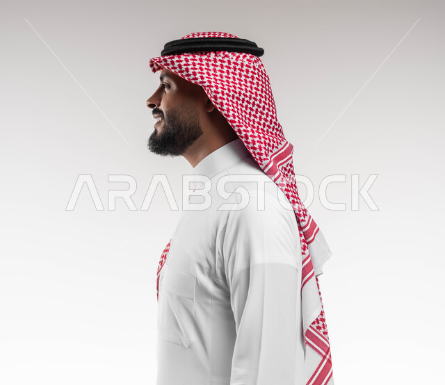 عرض ثياب الماركات الرجالية السعودية، الاهتمام والاعتناء بالمظهر الخارجي، بورتريه مقرب من الجانب لشاب عربي خليجي سعودي مبتسم يرتدي الشماغ والثوب التقليدي ينظر الى شيء ما بتعابير السرور والسعادة، التزين و التجمل و الترتيب, خلفية بيضاء