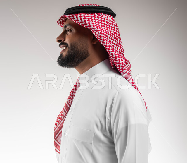 التجمل و الترتيب والتزين, بورتريه مقرب من الجانب لشاب عربي خليجي سعودي مبتسم يرتدي الشماغ والثوب التقليدي ينظر الى شيء ما بتعابير السرور والسعادة، عرض ثياب الماركات الرجالية السعودية، الاهتمام والاعتناء بالمظهر الخارجي، خلفية بيضاء