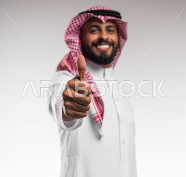 إشارات وحركات الاعجاب والرضا، بورتريه مقرب لشاب عربي خليجي سعودي مبتسم يرتدي الشماغ والثوب التقليدي يرفع إصبع الابهام الى الاعلى، مفهوم الإمتياز والتميز، إيماءات الموافقه والقبول، خلفية بيضاء