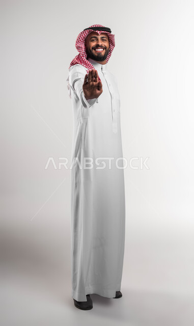 Non-acceptance and rejection of harm, portrait of a smiling Saudi Gulf Arab young man wearing a shemagh and traditional dress, raising his hand with movements of rejection, feeling fear and discomfort, asking for a ban and to stop doing something, full-length body portrait, white background