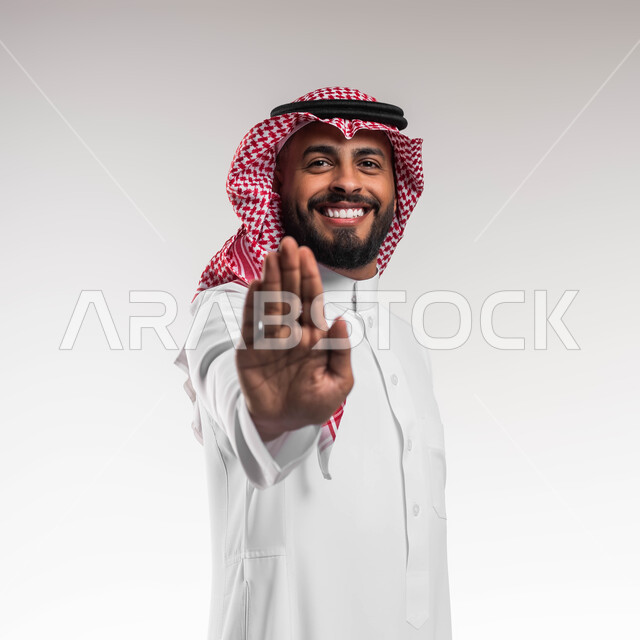عدم القبول ورفض الأذى, طلب المنع والتوقف عن فعل شيء ما، بورتريه مقرب لشاب عربي خليجي سعودي مبتسم يرتدي الشماغ والثوب التقليدي يرفع يده بحركات الرفض, الوقوف والنظر إلى الكاميرا، خلفية بيضاء