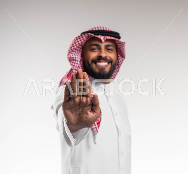 طلب المنع والتوقف عن فعل شيء ما، بورتريه مقرب لشاب عربي خليجي سعودي مبتسم يرتدي الشماغ والثوب التقليدي يرفع يده بحركات الرفض, الوقوف والنظر إلى الكاميرا, عدم القبول ورفض الأذى، خلفية بيضاء