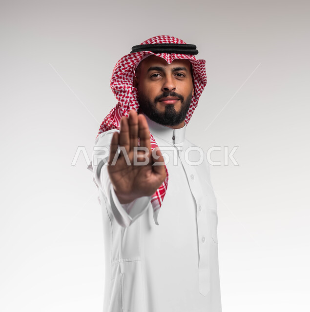 Gestures and expressions of seriousness and firmness, non-acceptance and refusal to harm, close-up portrait of a smiling Saudi Gulf Arab young man wearing a shemagh and traditional dress, raising his hand with movements of rejection, feeling fear and discomfort, asking for a ban and to stop doing something, white background.