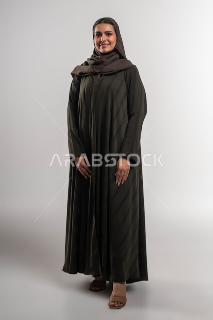 الاهتمام بالمظهر الخارجي, مفهوم الأنوثة والنعومة, الوقوف بابتسامة وثقة بالنفس, بورتريه لإمرأة عربية خليجية سعودية ترتدي العباءة والحجاب تنظر إلى الكاميرا بإيماءات السرور والسعادة, صورة الجسم بالطول الكامل, خلفية بيضاء