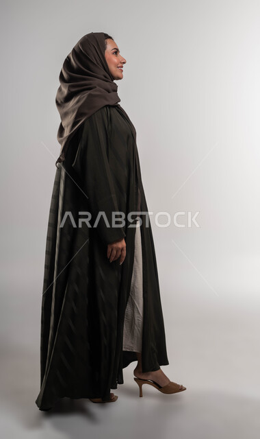 الوقوف بثقة بالنفس, الاهتمام بالمظهر الخارجي, مفهوم الأنوثة والنعومة, بورتريه من الجانب لإمرأة عربية خليجية سعودية ترتدي العباءة والحجاب تنظر إلى شيء ما بإيماءات السرور والسعادة, صورة الجسم بالطول الكامل, خلفية بيضاء