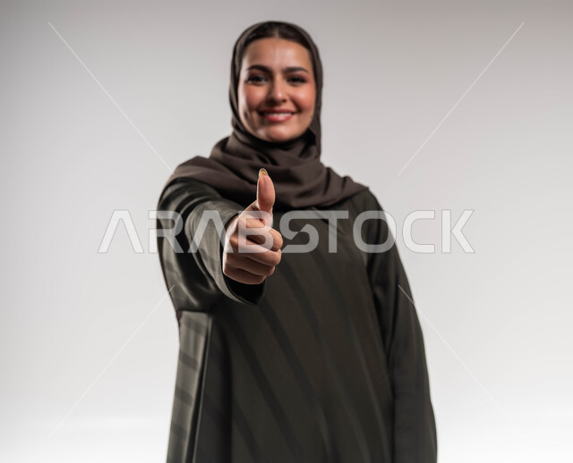 الرضا والإعجاب والقبول, بورتريه مقرب لإمرأة عربية خليجية سعودية مبتسمة ترتدي العباءة والحجاب ترفع يديها بإيماءات الجودة والامتياز, النجاح وتحقيق أهداف الشركة, مهن ووظائف نسائية سعودية, خلفية بيضاء