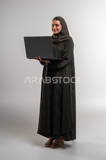 The concept of developing future projects and plans, Saudi women’s professions and jobs, business management through modern technologies and devices, a portrait of a smiling Saudi Gulf Arab woman wearing an abaya and hijab holding a laptop, smart and safe electronic marketing, full body image, white background