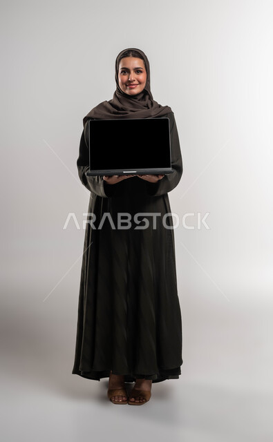 التطوير المهني عبر التطبيقات الحديثة لإدراة المشاريع, إنجاز ومتابعة مهام العمل عن بعد, استخدام الأجهزة والتقنيات الحديثة, بورتريه لسيدة أعمال عربية خليجية سعودية مبتسمة محجبة ترتدي العباءة السوداء تمسك الحاسب المحمول بشاشة سوداء فارغة, صورة الجسم بالكامل، خلفية بيضاء
