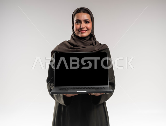 استخدام الأجهزة والتقنيات الحديثة, إنجاز ومتابعة مهام العمل عن بعد, بورتريه مقرب لسيدة أعمال عربية خليجية سعودية مبتسمة محجبة ترتدي العباءة السوداء تمسك الحاسب المحمول بشاشة سوداء فارغة, التطوير المهني عبر التطبيقات الحديثة لإدراة المشاريع, خلفية بيضاء