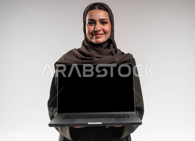 Completing and following up on work tasks remotely, using modern devices and technologies, a close-up portrait of a smiling, veiled Saudi Arabian Gulf businesswoman wearing a black abaya holding a laptop with a blank black screen, professional development through modern applications for project management, white background