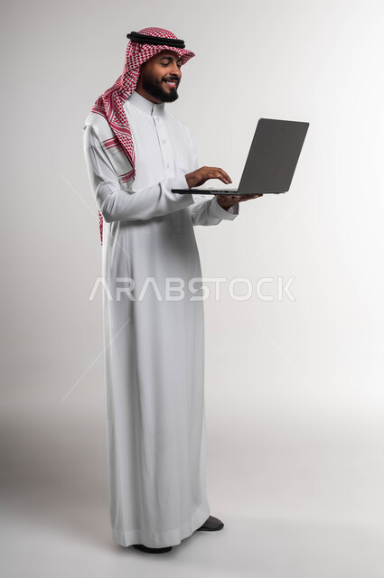 Saudi professions and jobs, integrating technology and developing a work plan and mechanism through modern technologies, completing office work, close-up portrait of a Saudi Gulf Arab employee wearing a traditional thobe and shemagh working on a laptop, following up on administrative affairs, full-length body photo, white background